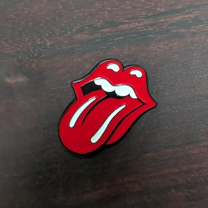 May include: Red and white enamel pin featuring the iconic Rolling Stones tongue logo. The tongue is sticking out with a white outline and a black line across the top.