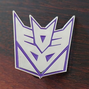 May include: A silver-colored metal pin with a purple outline in the shape of the Decepticon symbol from the Transformers franchise.