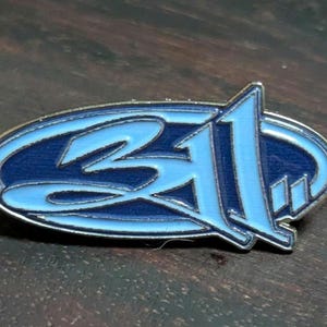 May include: A blue and silver enamel pin featuring the number 311 in a stylized font. The pin has a dark blue oval background with a light blue outline and lettering. The pin is set against a dark wood grain surface.