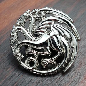 May include: A circular silver-toned metal pin featuring a three-headed dragon design. The dragon is depicted in profile with its wings spread and tails curled within the circular frame. The metallic surface has a polished, reflective finish.