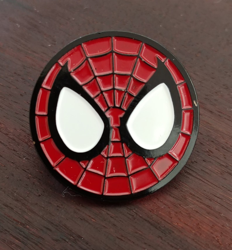 May include: A red and black enamel pin featuring a stylized image of Spider-Man's mask.
