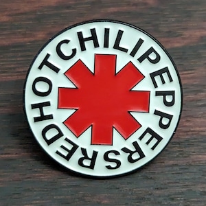 Può includere: Spilla con il logo della band Red Hot Chili Peppers su sfondo bianco e scritta nera. Il logo presenta una stella rossa.