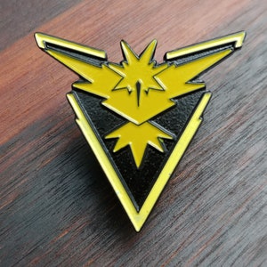 Op de afbeelding: Een zwarte en gele emaille pin met de iconische Pokémon-personage Articuno uit de Pokémon-franchise. De pin heeft de vorm van een driehoek met een gestileerd vogelontwerp.