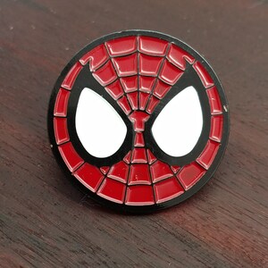 May include: A red and black enamel pin featuring a stylized image of Spider-Man's mask. The mask is white with black outlines and a red spider web pattern.