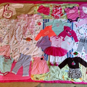 Pode incluir: Um cobertor de bebê rosa e branco com várias roupas de bebê aplicadas, incluindo macacões, camisas, vestidos e calças. O cobertor apresenta uma variedade de padrões e cores, incluindo rosa, branco, azul, verde e amarelo.