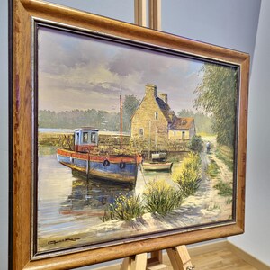May include: Oil painting of a coastal scene with a blue and red boat docked in a calm waterway. A stone building and a path are visible in the background. The painting is framed in a wooden frame and displayed on a wooden easel.