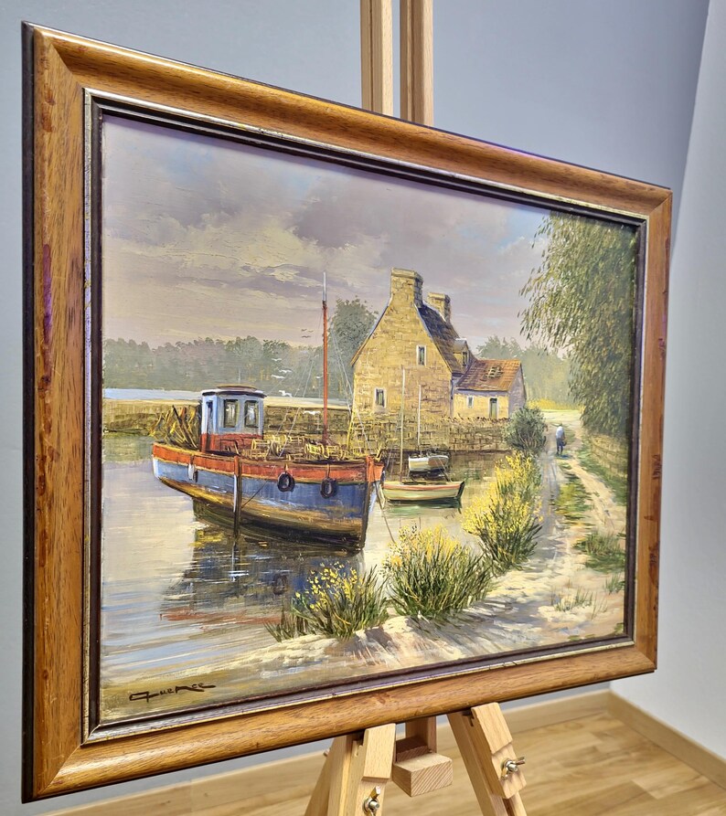 May include: Oil painting of a coastal scene with a blue and red boat docked in a calm waterway. A stone building and a path are visible in the background. The painting is framed in a wooden frame and displayed on a wooden easel.