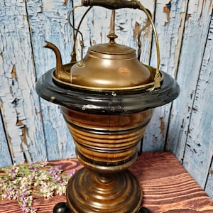 Op de afbeelding: Antiek houten standaard met een koperen ketel erop. De standaard heeft een zwart geverfde rand en een ronde basis met drie zwarte poten. De ketel heeft een houten handvat en een messing tuit.