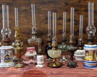 May include: A collection of antique oil lamps with glass chimneys. The lamps vary in size, shape, and color, with bases in white, brass, and painted ceramic. Some have floral designs, while others feature ornate metalwork.