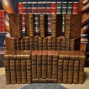 May include: A collection of antique books with brown leather covers and gold-colored decorative accents. The spines feature titles and ornate designs. The books are arranged in a stacked display, showcasing their aged appearance and historical charm.