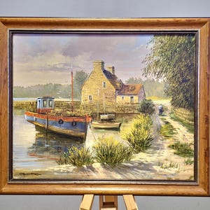 May include: Oil painting of a coastal scene with a blue and red boat docked near a stone building. The artwork is framed in a wooden frame and depicts a serene waterside landscape with a path and a person walking.