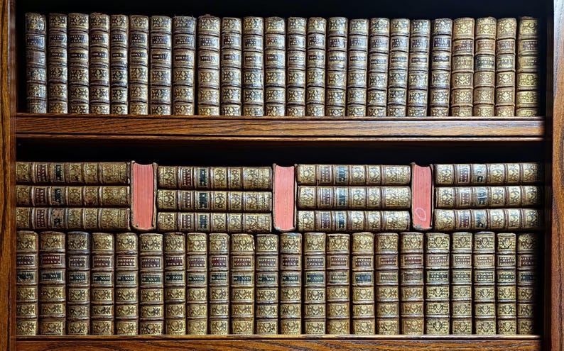 May include: A wooden bookshelf filled with antique books. The books have aged, textured covers with gold lettering and designs. A few books have a contrasting red spine, adding a pop of color to the display.