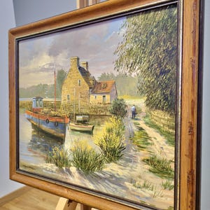 May include: Oil painting of a coastal scene with a blue and red boat docked near a stone building. A person walks along a path beside the water. The painting is in a wooden frame and displayed on an easel.