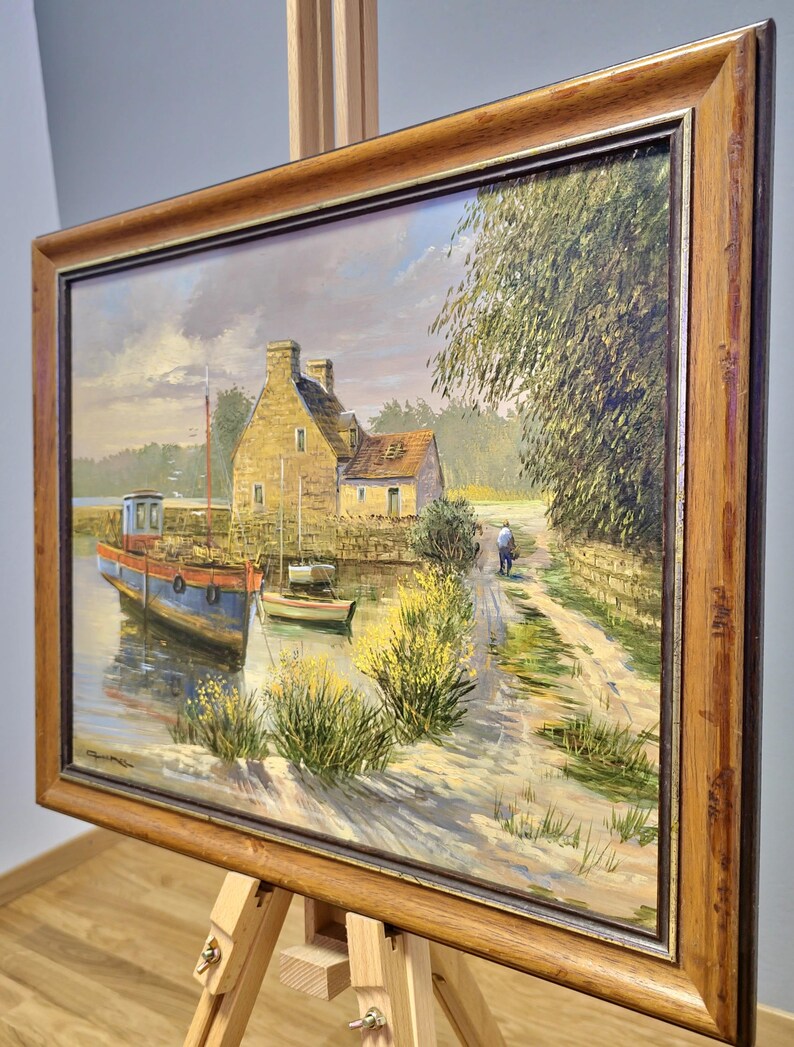 May include: Oil painting of a coastal scene with a blue and red boat docked near a stone building. A person walks along a path beside the water. The painting is in a wooden frame and displayed on an easel.