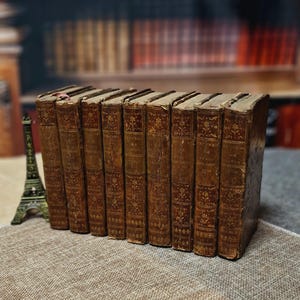 May include: A collection of antique books with brown leather covers. The spines feature embossed floral patterns and titles in gold lettering. The books are arranged in a row on a textured surface, with a miniature Eiffel Tower in the background.
