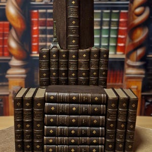 Può includere: Una collezione di libri antichi con copertine in pelle marrone scuro e dettagli dorati in rilievo. I libri sono impilati a forma di piramide, con una piccola statuetta della Torre Eiffel in cima. Lo sfondo mostra una biblioteca sfocata.