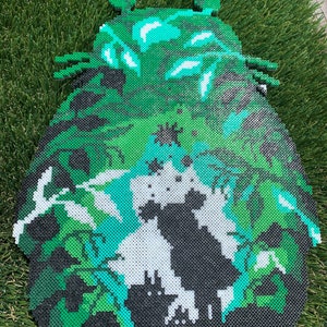 May include: A green, black, and white beaded art piece depicting a cartoon character. The character is a large, round creature with a large head and small ears. The character is surrounded by green leaves and vines.