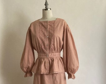 May include: A two-piece, light-brown outfit on a mannequin. The top has a square neckline, lace detailing, and button closures. The long sleeves are gathered at the cuffs. The skirt is a long, straight style with a ruffled hem. The outfit is made of a soft fabric.