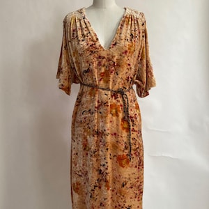 May include: A peach-colored dress with a V-neck and short sleeves. The dress features a floral pattern in shades of red, orange, and brown. A thin, dark brown belt cinches the waist. The dress is displayed on a mannequin.