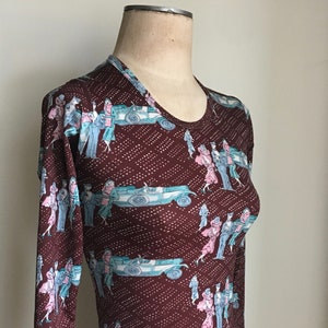 Puede incluir: Una camisa marrón de manga larga con un patrón repetido de coches antiguos y personas en un estilo de los años 20. La camisa tiene cuello redondo y una silueta ajustada.