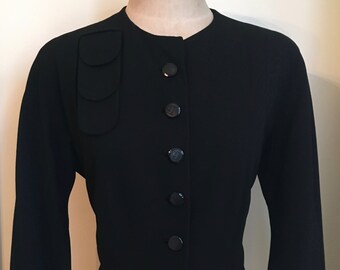 May include: A black fitted jacket with a round neckline and a row of black buttons down the front. The jacket has a unique detail on the shoulder with three overlapping fabric pieces.