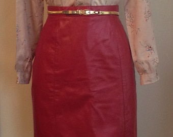 May include: A light pink blouse with a colourful abstract print and long sleeves. The blouse is worn with a red leather skirt and a gold belt.