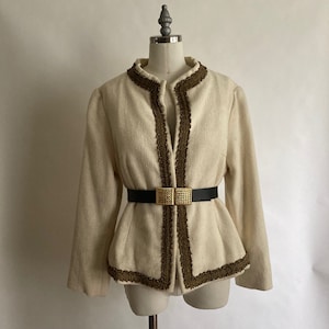 May include: A cream-coloured jacket with brown trim and a black belt with a gold buckle. The jacket has a V-neck and long sleeves.