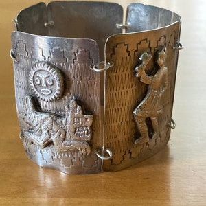 May include: A silver bracelet with a geometric design and three raised figures. The figures include a sun with a face, a dog, and a person holding a staff.