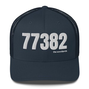 Może przedstawiać: Czapka trucker w kolorze granatowym z siatką w kolorze białym z tyłu. Z przodu czapki znajduje się liczba "77382" w kolorze białym i napis "the woodlands" w mniejszym rozmiarze w kolorze białym pod liczbą.