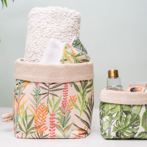 May include: Two fabric storage baskets with a tropical leaf print. The larger basket contains a rolled white towel and several white and patterned items. The smaller basket holds a clear bottle and a small packet. The baskets have a neutral-coloured rolled top.