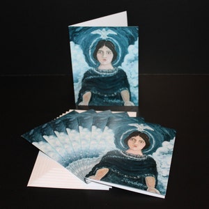 May include: A stack of greeting cards with a blue and white illustration of a woman with a dove above her head. The woman is wearing a dark blue robe and has a halo around her head. The text "J. Gordon" is visible in the bottom right corner of the card.