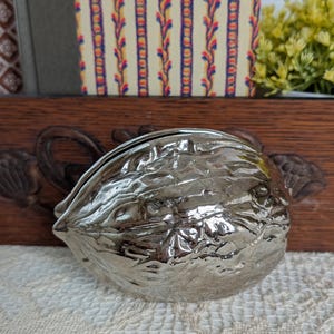 May include: A shiny, silver-toned metal object shaped like a walnut shell. The surface features a highly reflective, textured finish that mimics the natural, irregular ridges and crevices found on the exterior of a real walnut, with a visible seam.