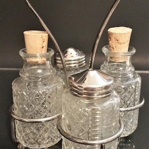 Könnte beinhalten: Ein silberner Metallständer mit vier gläsernen Salz- und Pfefferstreuern. Die Streuer haben einen klaren Glaskörper mit einem Diamantenmuster und Korkstopfen.