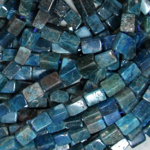 May include: A close-up of a collection of blue-green apatite beads. The beads are rectangular with rounded edges and are strung together on a blue cord. The beads have a polished, slightly translucent appearance.