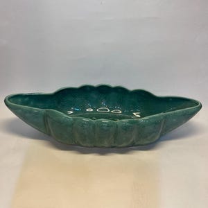 May include: A teal-colored, oblong ceramic planter with a scalloped rim. The planter has a glossy finish and a series of vertical ridges along the sides. The interior of the planter is smooth and reflects light.