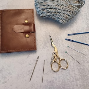 May include: A brown leather case with a buckle, a ball of grey yarn, gold scissors, blue knitting needles, and sewing needles are arranged on a light grey surface. Crafting and sewing supplies.