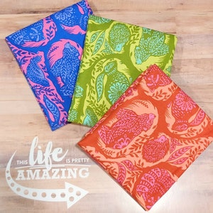 May include: Three fabric squares with a repeating pattern of animals and leaves. The fabric squares are blue, green, and orange. The text "This life is pretty amazing" is printed on the wooden surface below the fabric squares.