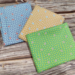 May include: Three folded fabric squares with geometric triangle patterns in blue, yellow, and green. The blue fabric has red triangles, the yellow has pink, and the green has purple. The fabrics are on a wooden surface.