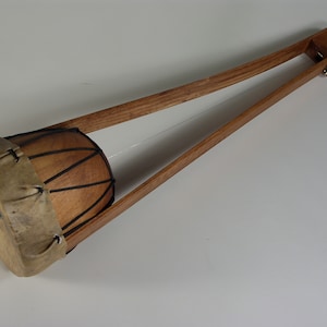 May include: A wooden musical instrument with a leather-covered drum at one end and a wooden tuning peg at the other end.