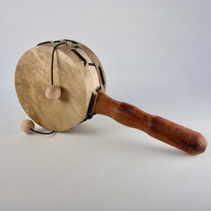 Op de afbeelding: Een houten handtrommel met een bruine handgreep en een leren trommelvel. Twee kleine houten balletjes zijn met touwtjes aan het trommelvel bevestigd.