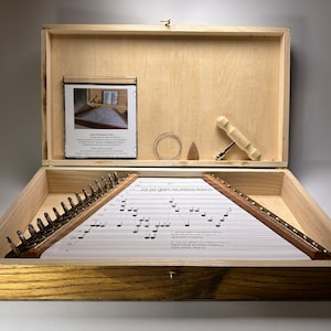 Può includere: Scatola di legno aperta contenente uno strumento musicale con corde e uno spartito. La scatola rivela i componenti dello strumento, tra cui una chiave di accordatura e corde di ricambio. Il telaio dello strumento è in legno.