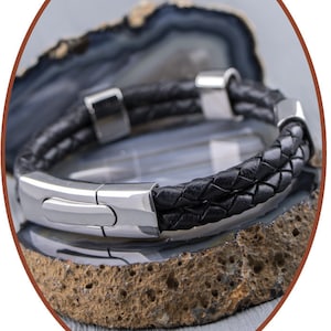 K&ouml;nnte beinhalten: Ein schwarzes Lederarmband mit einem silbernen Verschluss. Das Armband ist geflochten und hat eine polierte Metalloberfl&auml;che.