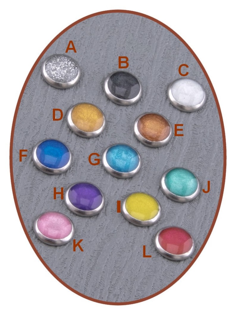 Op de afbeelding: Twaalf ronde, metalen, decoratieve knoppen met verschillende gekleurde epoxyharsinzetstukken. De knoppen zijn in een cirkel op een grijze achtergrond gerangschikt. De kleuren omvatten zilver, zwart, wit, goud, blauw, turquoise, paars, geel, roze en rood.