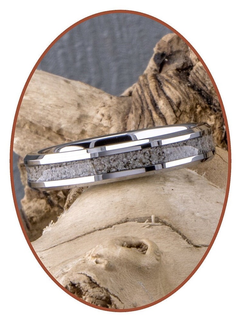 Op de afbeelding: Een zilveren ring met een band van witte geweininleg. De ring ligt op een stuk drijfhout.