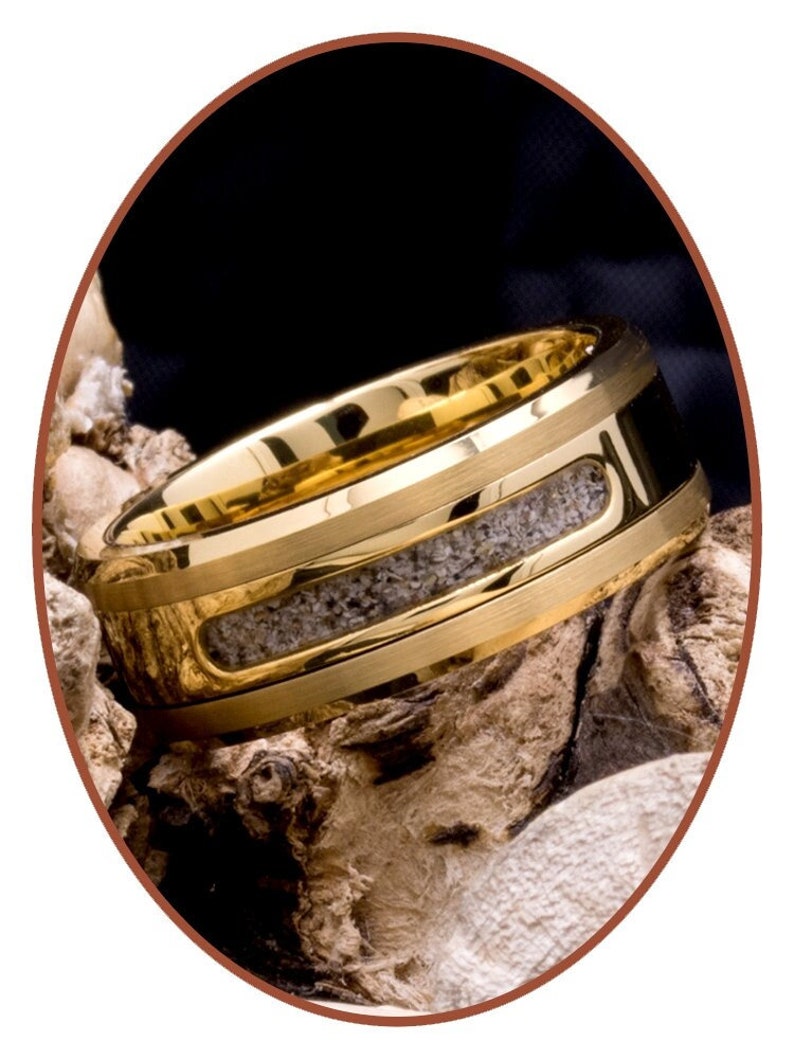 May include: A gold tungsten carbide ring with a band of grey inlay. The ring is on a piece of driftwood.