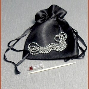 May include: A black satin pouch with a silver chain inside. The pouch is tied with a black ribbon. A small clear vial and a wooden toothpick are also included.