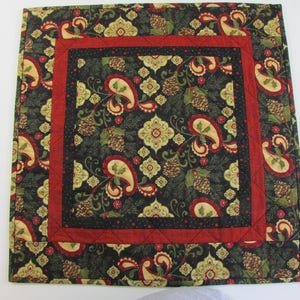 May include: A square quilted table runner with a black background and a paisley pattern in shades of green, red, and gold. The runner has a red and black border.