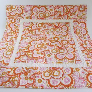 May include: A quilted bedspread with a floral pattern in shades of orange, pink, and white. The design features a large central square bordered by a white band. The overall aesthetic is vintage and decorative.