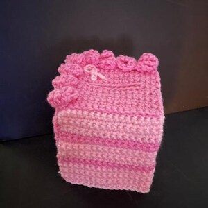May include: A handmade, pink crocheted item, possibly a cozy or cover. The piece features varying shades of pink, with a decorative top edge and a small pink bow. The item is likely a handmade craft.