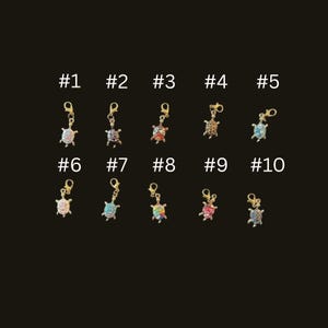 May include: A collection of ten small, decorative turtle charms with gold-colored clasps. Each charm is embellished with colorful rhinestones. The charms are numbered from one to ten against a dark background.